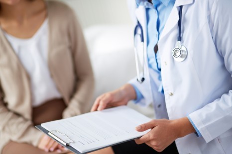 Patient in a doctors office with a doctor standing in front holding a clipboard